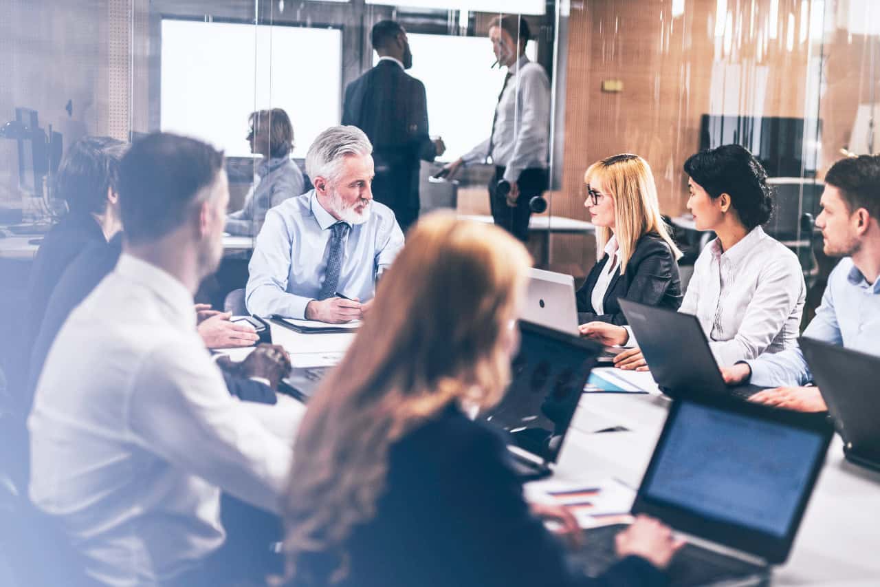 A professional leader with gray hair gestures while speaking to colleagues, appearing to be a manager or mentor in a business discussion.