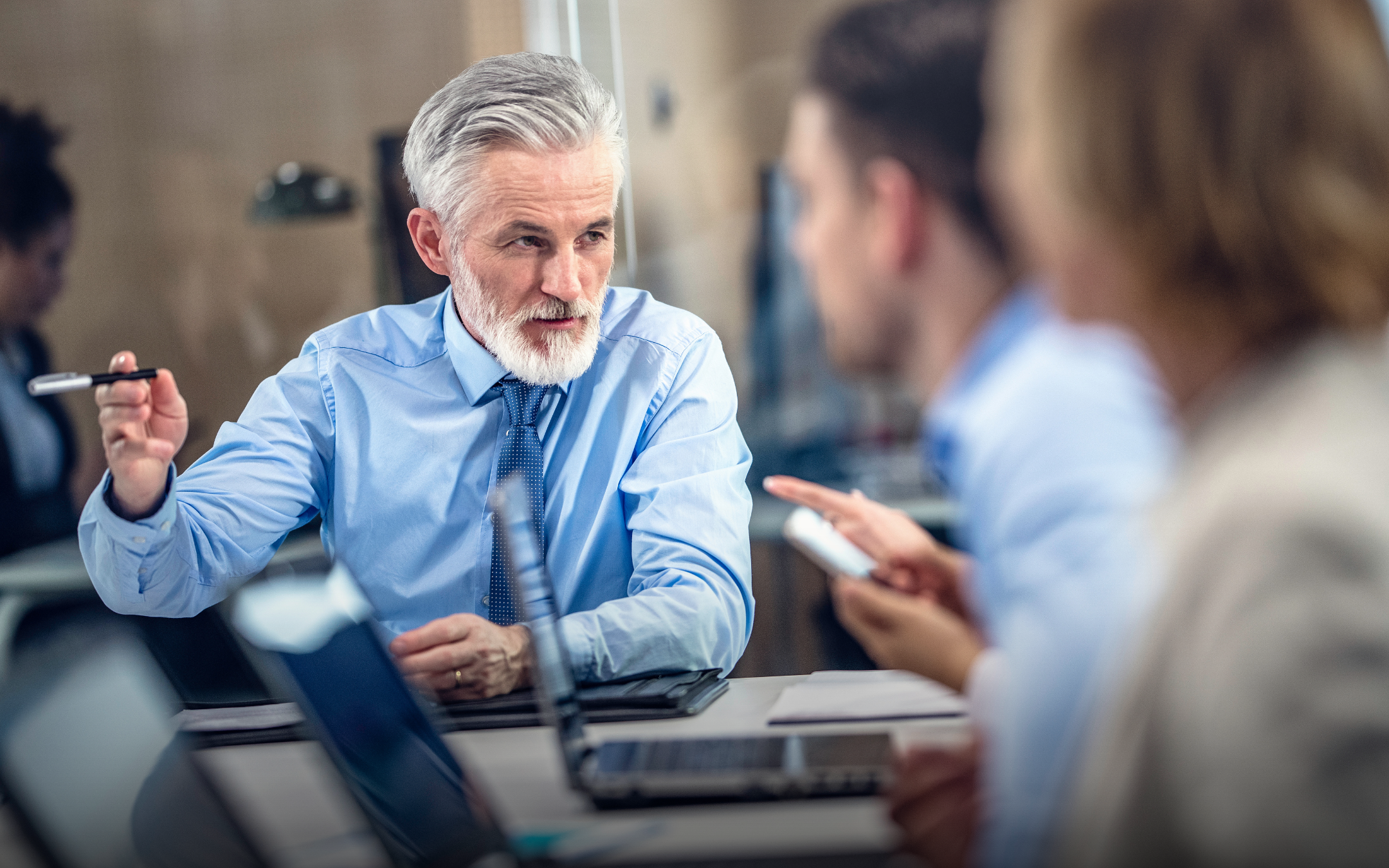 A professional leader with gray hair gestures while speaking to colleagues, appearing to be a manager or mentor in a business discussion.